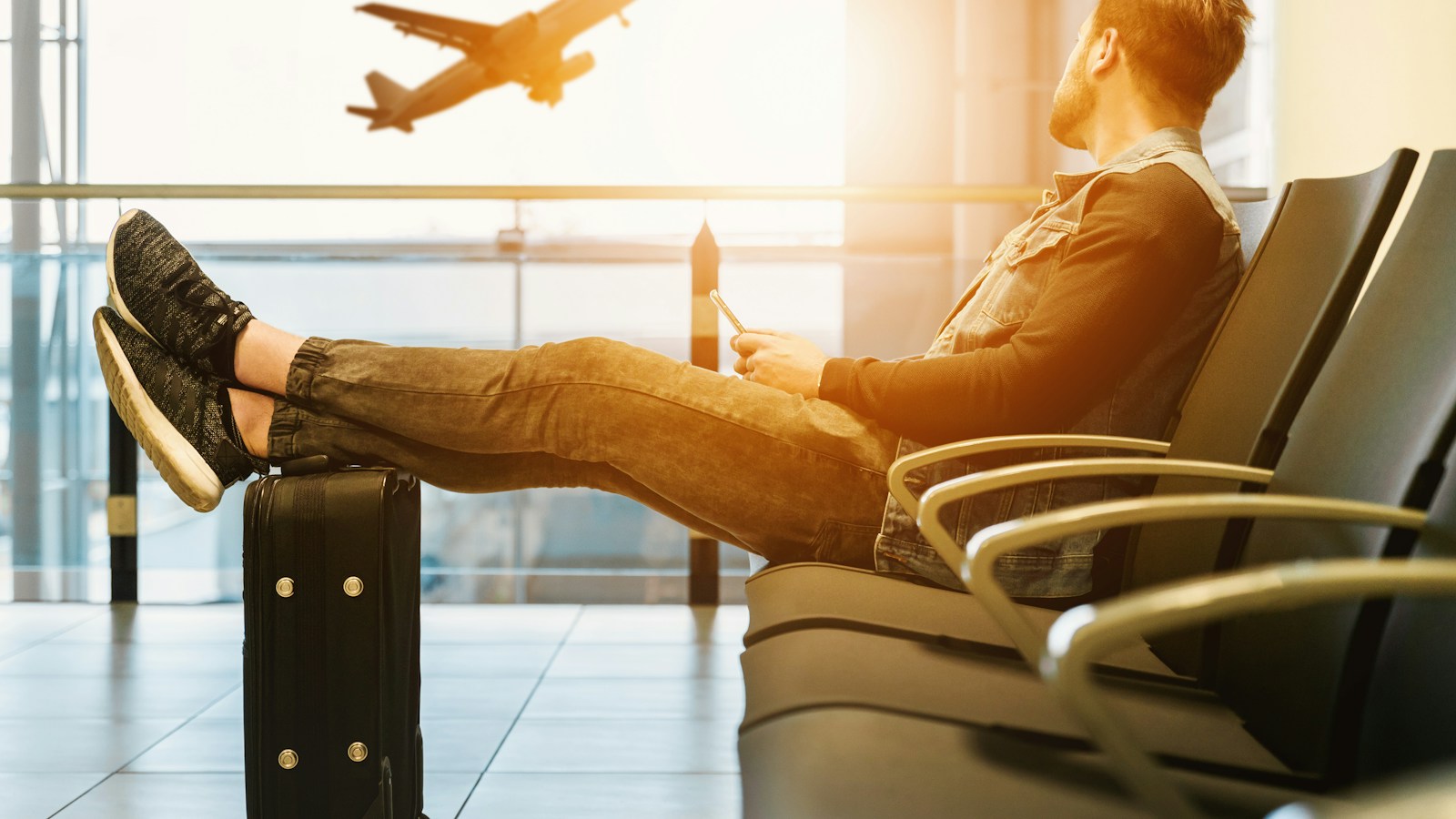 Viajante descansando no aeroporto com os pés sobre a mala, olhando um avião decolar