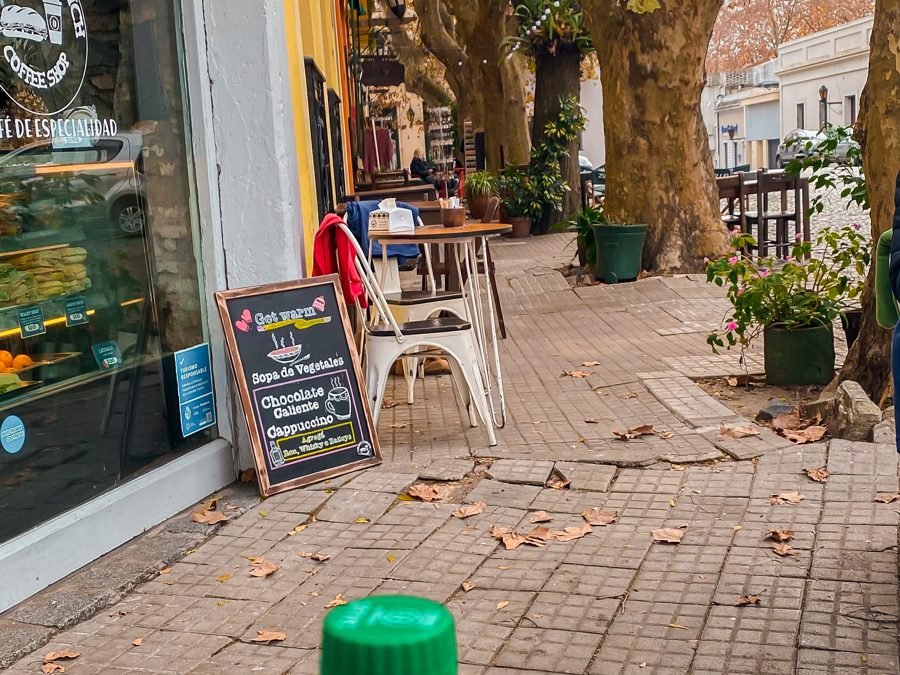Mesa de café na calçada, com árvores de outono e casas coloridas ao fundo, em Colônia do Sacramento