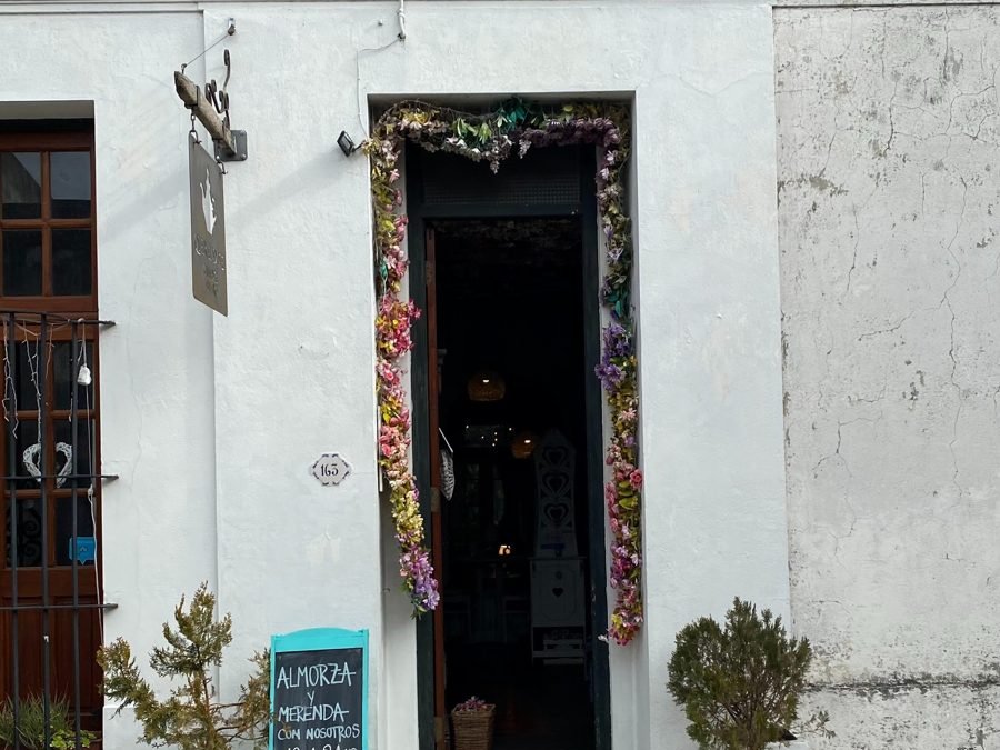 Fachada branca de uma casa de chá com porta enfeitada de flores e lousa anunciando almoço e lanche