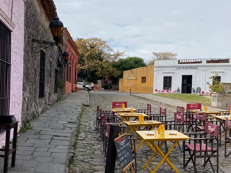 Mesas amarelas de restaurante sobre a rua de pedra, com lousa de câmbio na frente