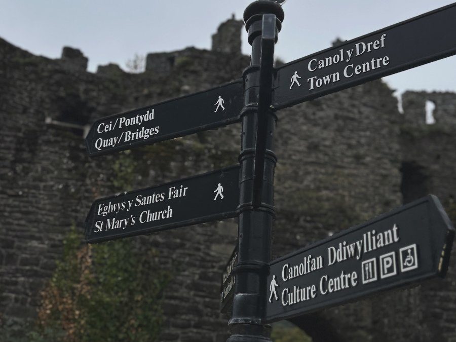 Placa de direção bilíngue em galês e inglês diante da muralha medieval de Conwy