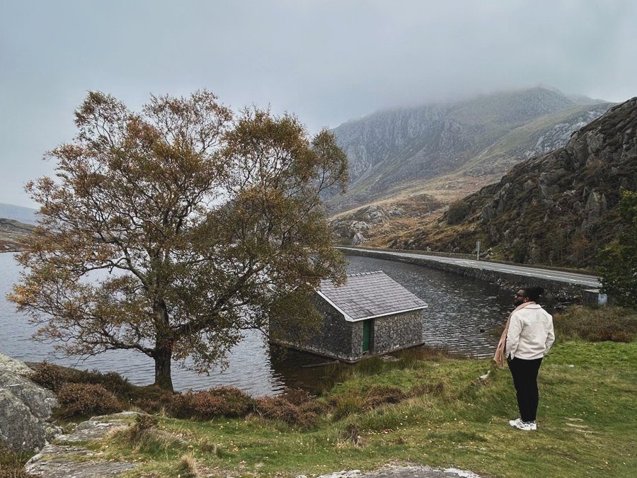 JP na beira do Llyn Ogwen, com a casinha de pedra, uma árvore de outono e montanhas na neblina