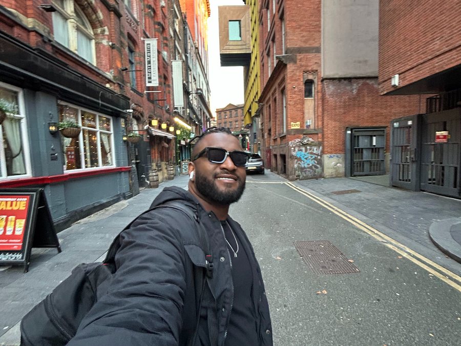 José Pedro sorrindo numa selfie de mochila nas costas, numa rua de prédios de tijolo