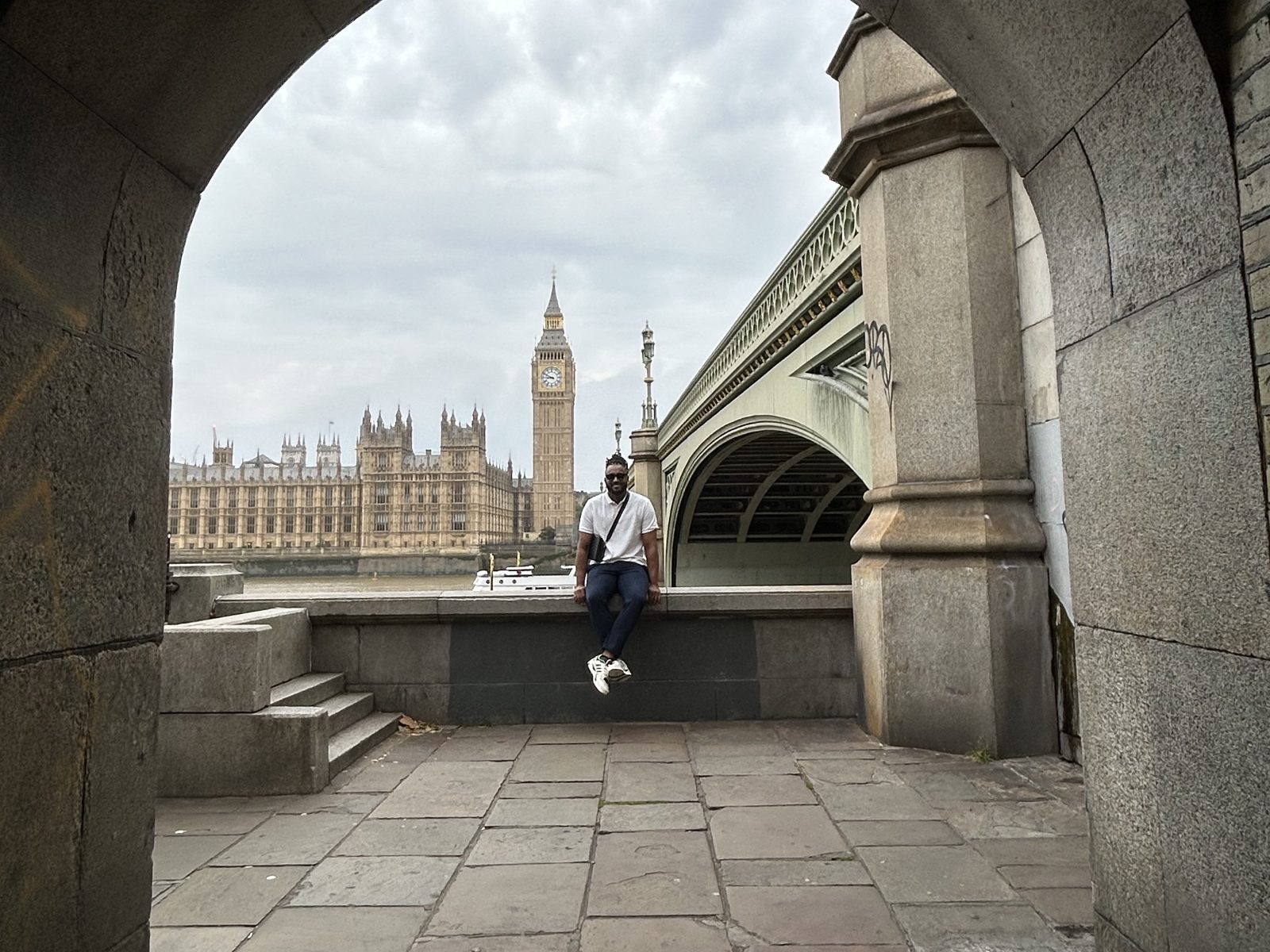 JP sentado na mureta à beira do Tâmisa, com o Big Ben e o Parlamento emoldurados por um arco sob a Westminster Bridge