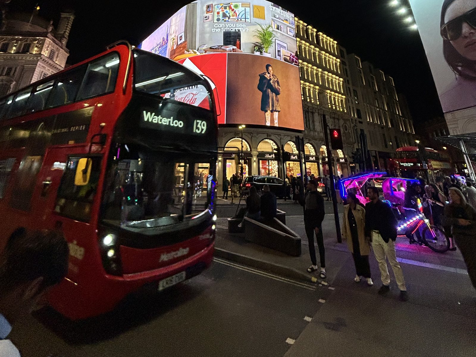 Ônibus vermelho de dois andares passando pelos telões iluminados de Piccadilly Circus à noite