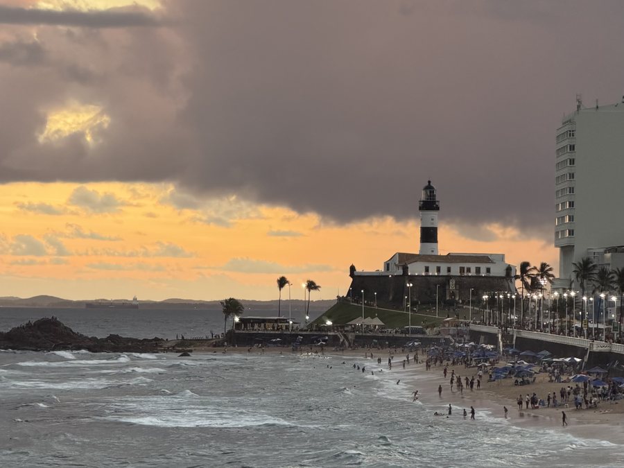Farol da Barra ao entardecer sob nuvens de tempestade, com a praia cheia e o mar agitado em Salvador