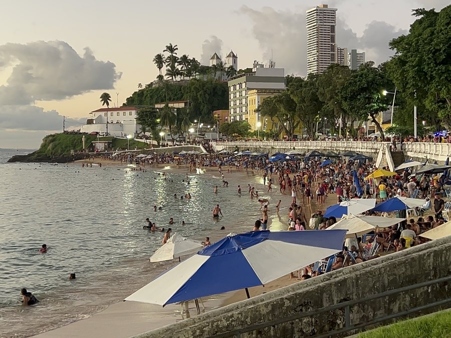 Porto da Barra lotado no fim de tarde, com guarda-sóis azuis e brancos e o mar calmo